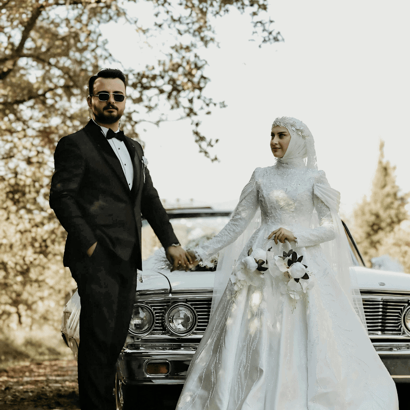 Wedding couple in formal attire holding hands in front of a vintage car outdoors.