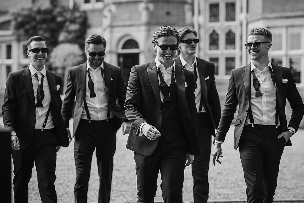 .Groom and groomsmen walking in tuxedos; pre-ceremony groom prep style; groomsmen wearing sunglasses; relaxed wedding photography Essex.