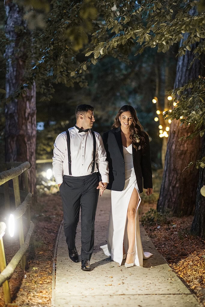Elegant couple walking hand in hand through a wooded path at night, illuminated by soft lighting. Captured by award-winning wedding photographer Scott Miller